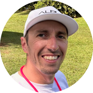 A man wearing a white cap and shirt is smiling at the camera in an outdoor setting with green grass and trees in the background.