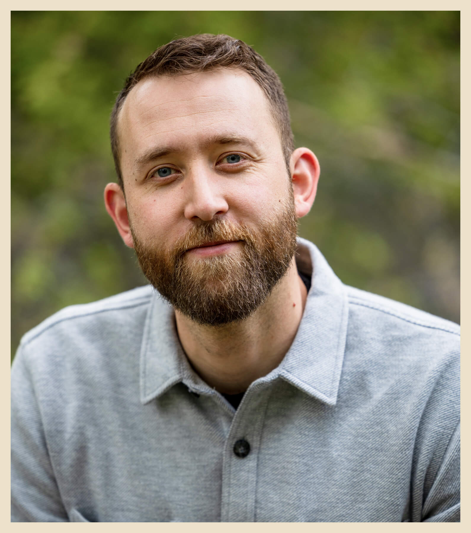 A person with a beard and short hair wearing a light gray collared shirt, standing outdoors with a blurred green background.