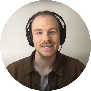 A person with short hair wearing a brown jacket and headphones, smiling at the camera against a white background.