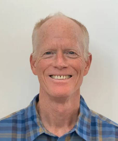 A smiling man with short, light-colored hair, wearing a blue plaid shirt, stands against a plain background.