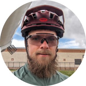 A man with a beard wearing a maroon bicycle helmet, clear safety glasses, and a green shirt stands outside with a building in the background.